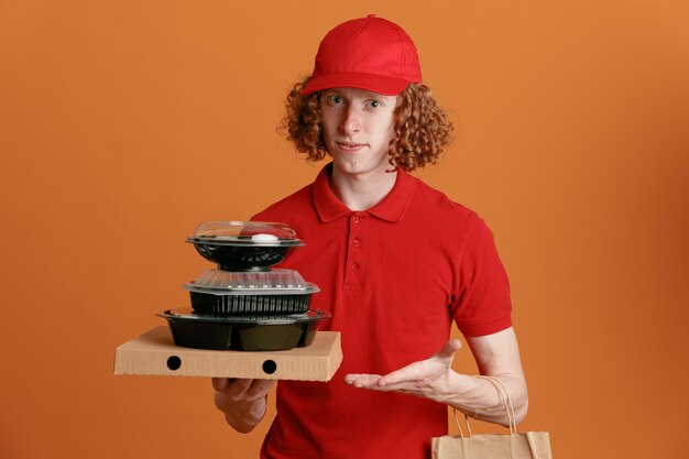 Bezorger werknemer in rode dop leeg t-shirt uniform met pizzadoos voedselcontainers met papieren zak presenteren met arm van hand die zelfverzekerd over oranje achtergrond kijkt