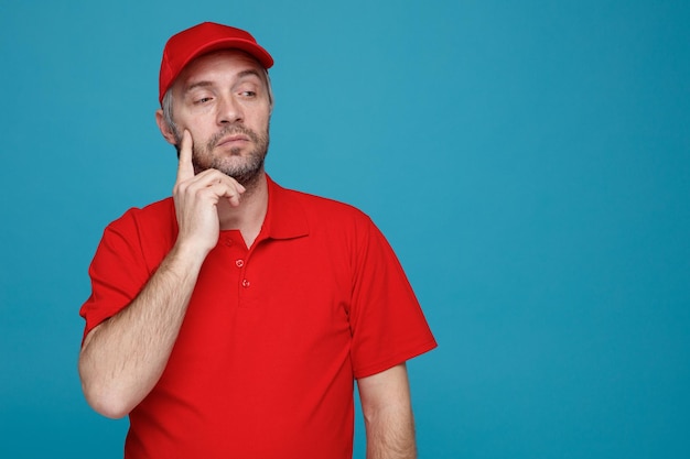 Bezorger werknemer in rode dop leeg t-shirt uniform opzij kijkend met peinzende uitdrukking denkend over blauwe achtergrond