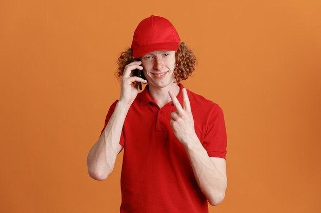 Bezorger werknemer in rode dop leeg t-shirt uniform praten op mobiele telefoon kijken camera glimlachen met nummer twee of vsign staande over oranje achtergrond