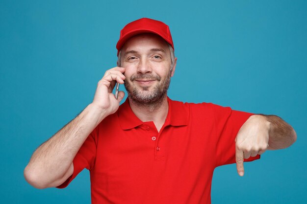Bezorger werknemer in rode dop leeg t-shirt uniform praten op mobiele telefoon wijzend met wijsvinger naar beneden glimlachend kijken naar camera staande over blauwe achtergrond