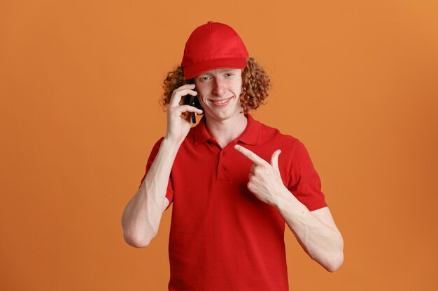 Bezorger werknemer in rode dop leeg tshirt uniform praten op mobiele telefoon wijzend met wijsvinger kijken camera glimlachend vrolijk staande over oranje achtergrond