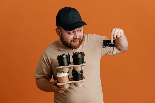 Bezorger werknemer in zwarte pet en blanco t-shirt uniform met koffiekopjes met creditcard blij en positief glimlachend vrolijk staande over oranje achtergrond