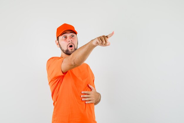 Bezorger wijst naar iets weg in oranje t-shirt, pet en kijkt verrast. vooraanzicht.