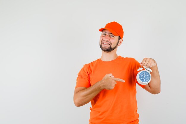 Bezorger wijzend op wekker in oranje t-shirt, pet en vrolijk kijkend. vooraanzicht.