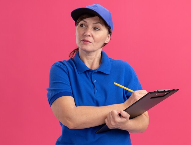 Bezorgingsvrouw van middelbare leeftijd in blauw uniform en GLB-holdingsklembord en potlood die opzij met ernstige zekere uitdrukking kijken die zich over roze muur bevindt