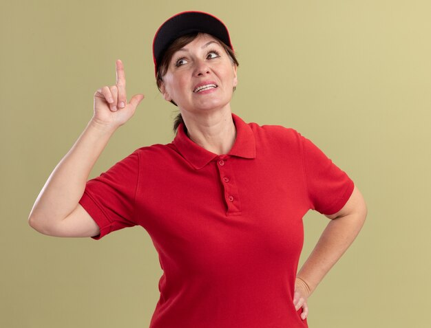 Bezorgingsvrouw van middelbare leeftijd in rood uniform en GLB die voorzijde glimlachend zelfverzekerd tonen die wijsvinger bekijken die nieuw groot idee hebben die zich over groene muur bevinden