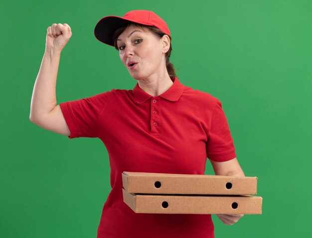 Bezorgingsvrouw van middelbare leeftijd in rood uniform en pet die pizzadozen bekijken die voorzijde opheffen vuist gelukkige en zekere status over groene muur opheffen