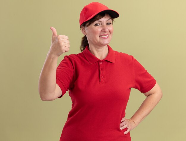 Bezorgingsvrouw van middelbare leeftijd in rood uniform en pet die voorzijde glimlachend vrolijk tonen die duimen tonen die zich over groene muur bevinden