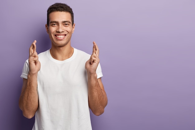 Biddende mannelijke student steekt gekruiste vingers op, vraagt om succes en geluk, glimlacht positief, bidt voordat hij een belangrijk examen in zijn leven haalt, draagt een casual wit t-shirt, poseert over de violette muur