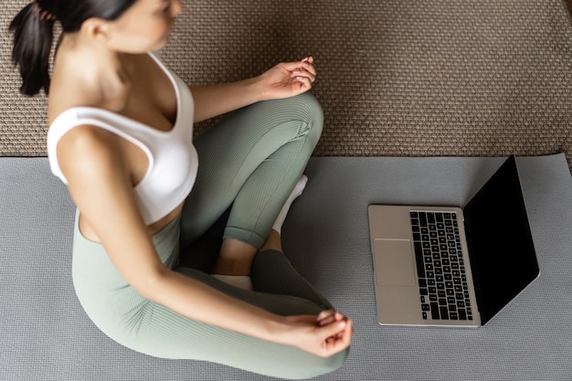 Bijgesneden opname van aziatische vrouw die thuis in vloermat mediteert, naar meditatiepodcast op laptop luistert, yoga online courase beoefent, activewear draagt.