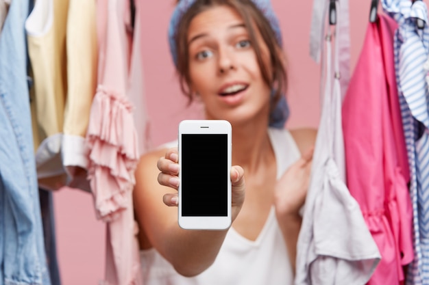 Bijgesneden opname van mooie vrouw met mobiele telefoon met leeg scherm terwijl je in de buurt van rek met kleren, laat zien welke kleren ze kiest in de online winkel. Kleding online kopen. Winkelen concept