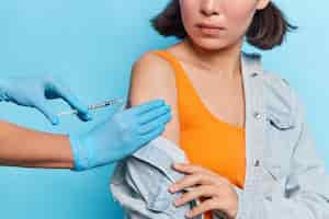 Gratis foto bijgesneden opname van onbekende vrouw maakt coronavirusvaccin om zichzelf te beschermen, draagt oranje t-shirt en spijkerblouse poseert tegen blauwe muur