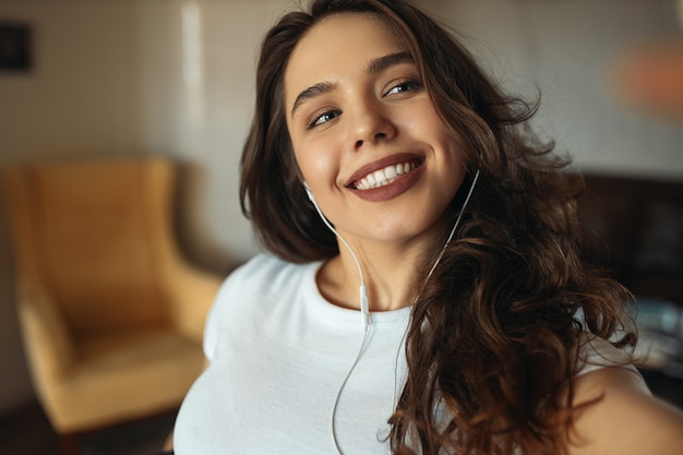 Bijgesneden portret van mooie jonge Europese vrouw met lang krullend haar en bruine lippenstift die oortelefoons draagt die met brede gelukkige glimlach kijken