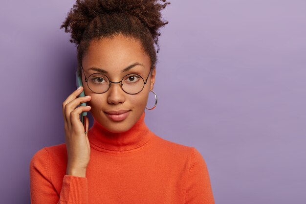 Bijgesneden schot van mooie donkere vrouw heeft ernstige uitdrukking, draagt een ronde bril en coltrui, heeft een telefoongesprek, krijgt een interessante suggestie