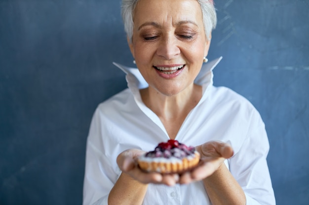 Bijgesneden schot van vrolijke aantrekkelijke grootmoeder in wit overhemd stuk versgebakken bessen taart houden voor verjaardag, met vrolijke gelaatsuitdrukking, breed glimlachend.