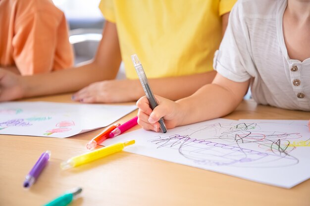 Bijgesneden weergave van kinderen schilderen op papier met pennen. Drie onherkenbare kinderen die aan tafel zitten en krabbels tekenen. Selectieve aandacht. Jeugd, creativiteit en weekendconcept