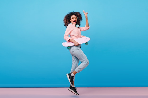 Binnen portret van gemiddelde lengte van zeker Afrikaans meisje in het roze skateboard van de overhemdsholding. Enthousiaste zwarte vrouw met krullend kapsel poseren met blauw interieur.