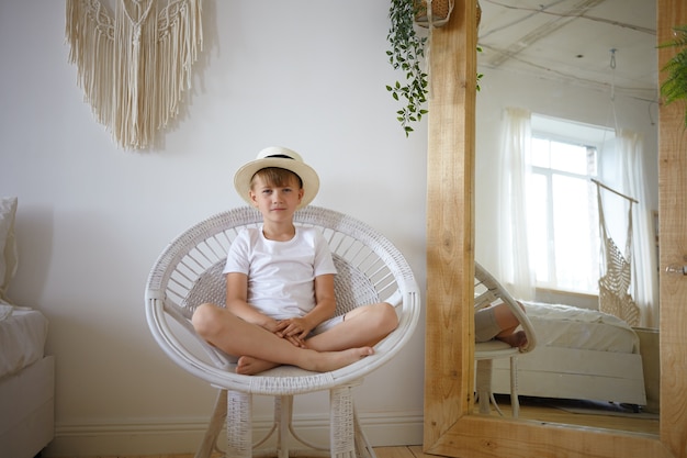 Binnen schot van 10-jarige jongen zitten in ronde fauteuil benen gekruist houden, kijken en glimlachen naar de camera, wit t-shirt en zomerhoed dragen. schattige mannelijke jongen poseren in slaapkamer met grote spiegel