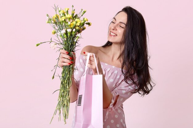Binnen schot van aangenaam ogende jonge vrouw glimlacht zachtjes, gekleed modieuze jurk, houdt cadeauzakje en bloemen, poses op lichtroze, verrukt. Vrouwendag concept