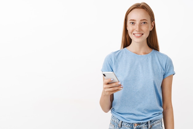 Binnen schot van aangename, vrolijke en vriendelijke roodharige vrouw met sproeten in blauw casual t-shirt met smartphone en glimlachend beleefd telefoonnummer opschrijven staande over grijze muur