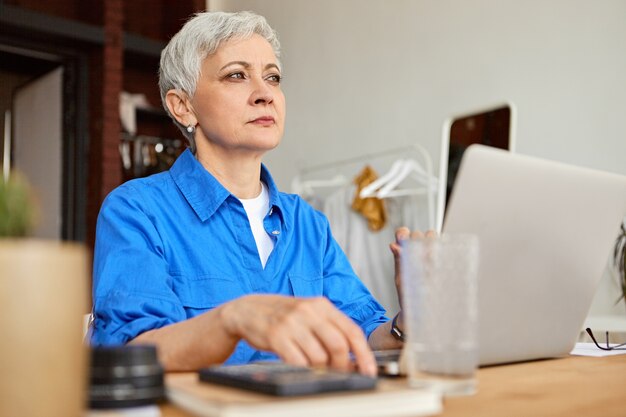 Binnen schot van aantrekkelijke grijze harige vrouwelijke gepensioneerde m / v werken als freelancer met behulp van laptopcomputer, om thuis te zitten bureau. Veroudering, pensioen, technologie, vrije tijd en beroep concept