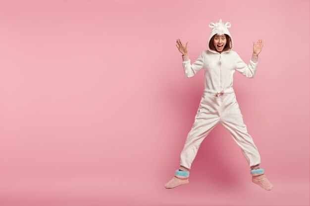 Binnen schot van dolblij gelukkig meisje in comfortabel huiselijk kigurumi kostuum, springt over roze ruimte