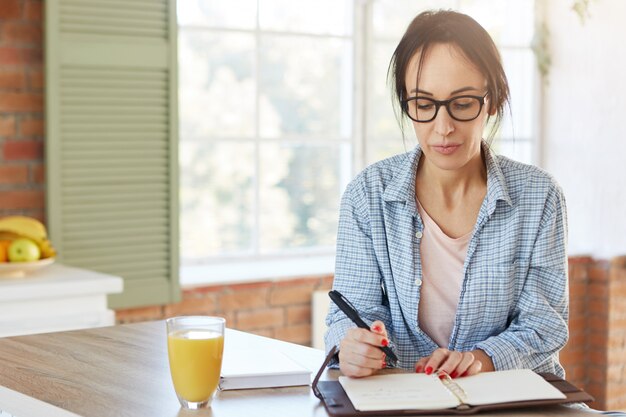 Binnen schot van ernstige Europese vrouw houdt pen vast, schrijft informatie die ze via internet heeft gevonden of boek in notitieblok, zit aan houten keukentafel met sap.