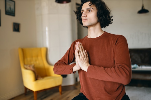 Binnen schot van geconcentreerde jonge man in vrijetijdskleding hand in hand samengedrukt tijdens het mediteren thuis tijdens yoga beoefening, de aandacht gericht op positieve gedachten.