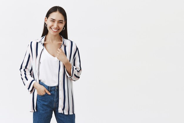 Binnen schot van gelukkige succesvolle jonge vrouwelijke ondernemer in gestreepte blouse en spijkerbroek, hand in de zak, vreugdevol glimlachend, tevreden na het openen van een eigen bedrijf over grijze muur