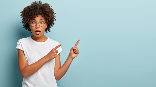 Binnen schot van onder de indruk verrast mooie vrouw met krullend haar, donkere huid, wijst beide vingers naar de rechterbovenhoek, geïsoleerd over blauwe muur, geschokt door hoge prijs op item in winkel