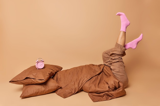 Gratis foto binnen schot van onherkenbare vrouw gekleed in pyjama houdt benen omhoog draagt sokken poses in bed onder deken wil niet opstaan geïsoleerd over beige studio achtergrond hard ontwaken