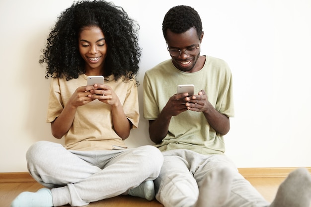 Binnen schot van terloops geklede jonge donkere familie zittend op witte lege muur op houten vloer met gadgets, met behulp van online apps