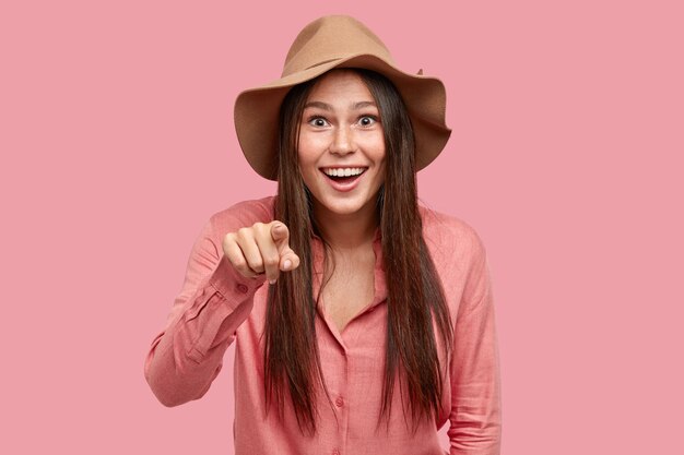 Binnen schot van vrolijke brunette vrouw met tevreden uitdrukking, glimlacht in grote lijnen, geeft direct in camera met wijsvinger