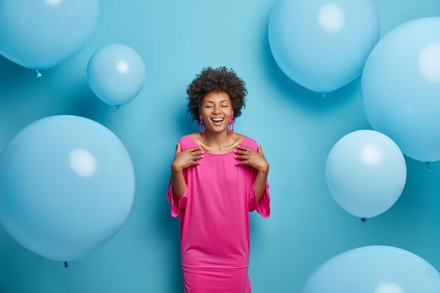 Binnen schot van vrolijke krullende vrouw in roze modieuze jurk, sluit de ogen, bereidt zich voor op een speciale gelegenheid, blij om gefeliciteerd met verjaardag te ontvangen, geïsoleerd op blauwe achtergrondgeluid, opgeblazen ballonnen