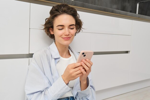 Binnenfoto van een gelukkig meisje met een smartphone die thuis op de vloer zit en mobiele telefoon gebruikt om video's te bekijken