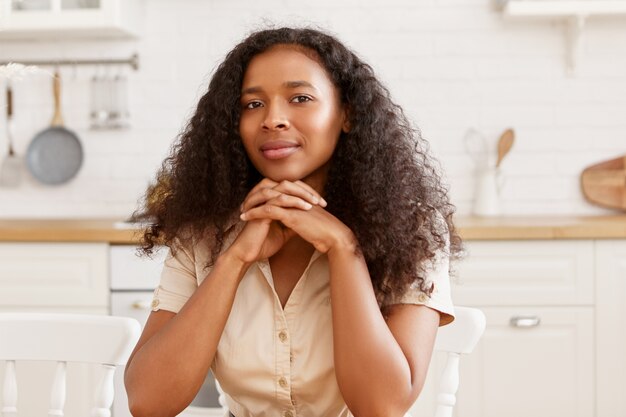 Binnenfoto van prachtige jonge zwarte donkere huid vrouw met Afro kapsel en gebruinde gebronsde huid koken in de keuken, zittend aan tafel, handen geklemd onder haar kin, kijken