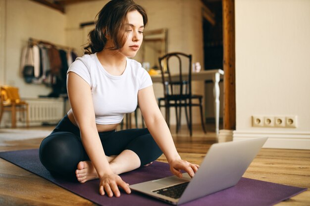 Binnenfoto van schattige plus size jonge vrouw zittend op de mat voor opengeklapte laptop, kijken naar online videozelfstudie door professionele fitnessinstructeur, trainen vanuit huis vanwege sociale afstand
