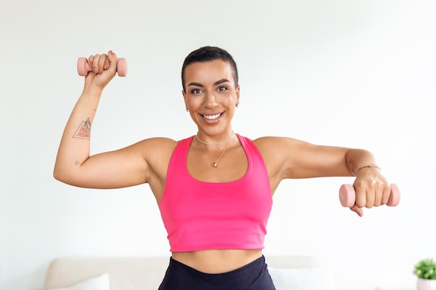 Binnenlandse training met gewichten positieve zwarte dame die oefeningen doet met halters die haar lichaam thuis versterken glimlachende jonge vrouw die aan haar biceps-spieren werkt en gezond blijft