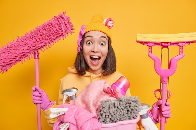 Gratis foto binnenopname van aziatische vrouw heeft een zeer verbaasd gezicht houdt de mond wijd open en merkt op dat de vuile kamer gaat schoonmaken alles houdt schoonmaakspullen staat bij de wasmand tegen de gele muur