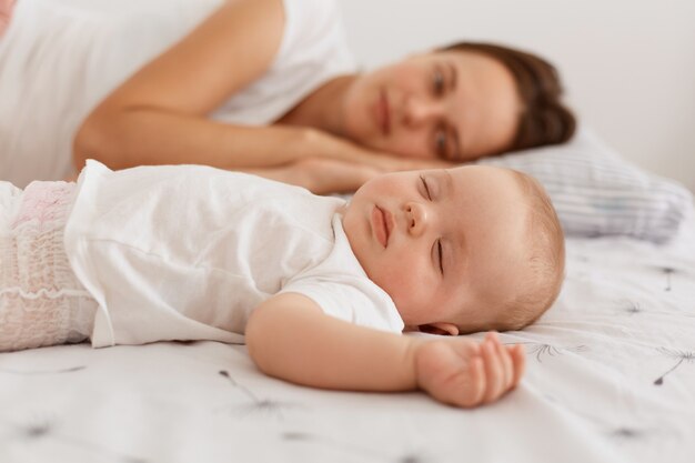 Binnenopname van een aantrekkelijke donkerharige vrouw met een wit t-shirt dat op bed ligt met haar baby-baby, die samen rust, mama die naar haar schattige dochter kijkt.