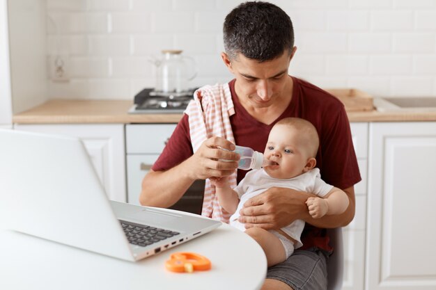 Binnenopname van een donkerharige knappe man met een casual t-shirt met een handdoek op zijn schouder, zittend aan tafel met laptop, babymeisje in handen houdend en water uit de fles gevend aan zijn dochter.