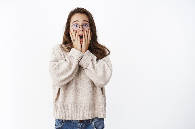 Binnenopname van een geschokte en verbaasde jonge vrouw met een bril en een trui die schreeuwde van schudde die de geopende mond met handen bedekte, bang en onzeker was, intens over de grijze muur staand.