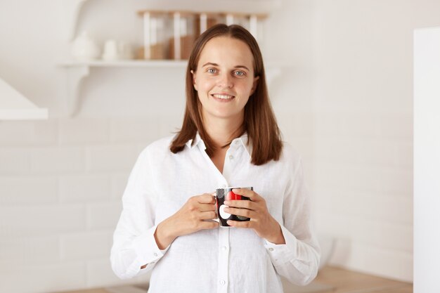 Binnenopname van een glimlachende mooie vrouw met een kopje koffie in de keuken, kijkend naar de camera met een vrolijke gezichtsuitdrukking, een vrouw met een wit casual stijlshirt.