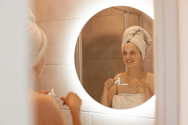 Binnenopname van een jonge volwassen vrouw die tanden poetst in de badkamer, naar haar reflectie in de spiegel kijkt, met blote schouders en een witte handdoek op haar haar staat. Gratis Foto