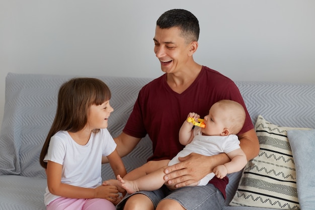 Binnenopname van gelukkige familie, glimlachende vader die met twee dochters op de bank speelt, schattige baby bijtspeelgoed, vader die tijd doorbrengt met charmante kinderen thuis.