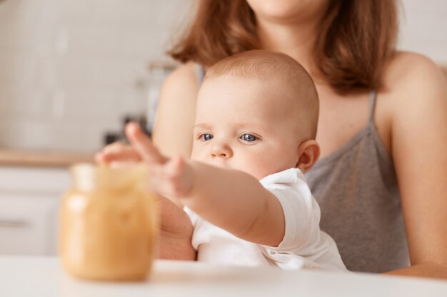 Binnenopname van gezichtsloze moeder die haar dochtertje met groentepuree voedt, charmante peuterbaby die hand uitrekt naar pot met eten, zich voedt.