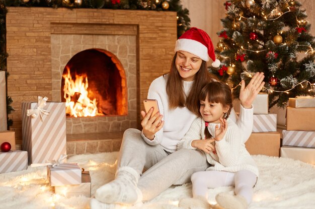 Binnenopname van moeder en dochtertje die videogesprek voeren of livestream uitzenden, handen zwaaien naar smartphonecamera, poseren in de buurt van open haard en kerstboom.