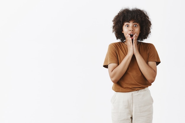 Binnenopname van onder de indruk sprakeloze knappe afrikaanse vrouw met krullend kapsel met handpalmen op gezicht, kaak laten vallen van verbazing staande met geopende mond opgewonden over grijze muur