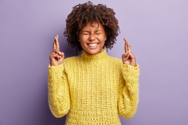 Binnenopname van positieve blije vrouw met donkere huid wenst het beste, kruist vingers, glimlacht breed, heeft ogen dicht, toont witte perfecte tanden draagt gele trui wacht op speciaal moment. Mogen dromen uitkomen