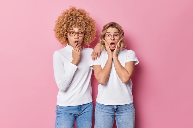 Binnenopname van twee bange jonge vrouwen staren emotioneel, voelen zich geschokt, dragen vrijetijdskleding kan niet geloven dat eigen ogen reageren op iets verbazingwekkends staren naar een camera geïsoleerd over roze achtergrond Omg concept